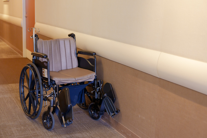 Empty wheelchair parked by cream wall on textured brown carpet suggests absence, evokes feelings of pause, awaiting return.