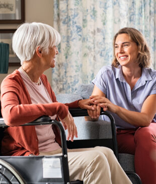 Mature woman comforting senior mom sitting on wheelchair at nursing home. Cheerful woman talking to old disabled mother in wheelchair at elder care centre. Loving caregiver taking care of elderly woman at home.